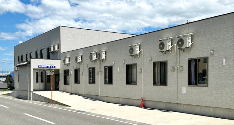 介護施設の建物外観。グレーのタイル張りモダン建築で、複数のエアコン室外機が設置されている。
