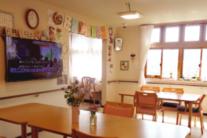 温かみのある共用リビング。テレビ、掲示板、テーブル、椅子が配置されている。